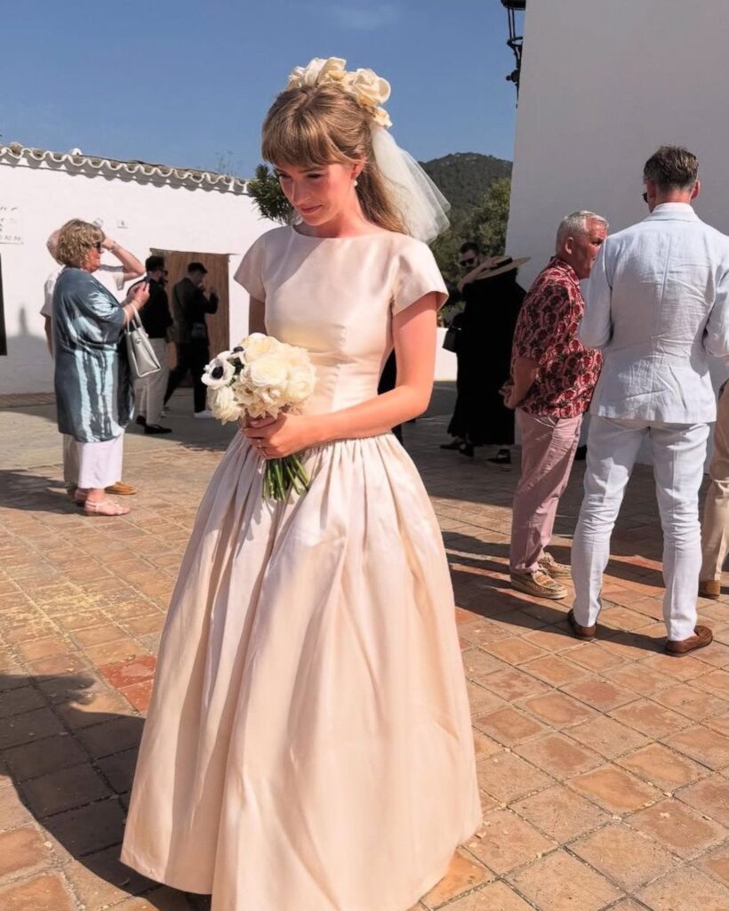 A bride in a cream satin wedding dress holding a white rose bouquet outdoors.