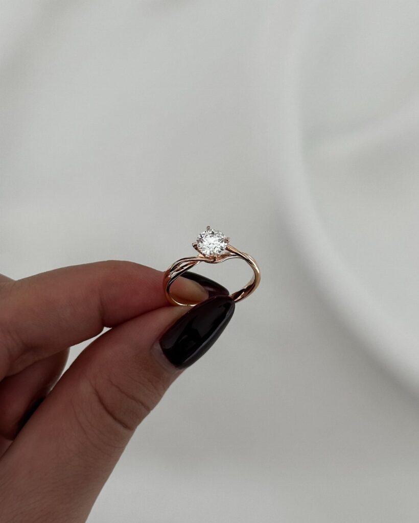 A rose gold diamond ring held by a hand with black nail polish.