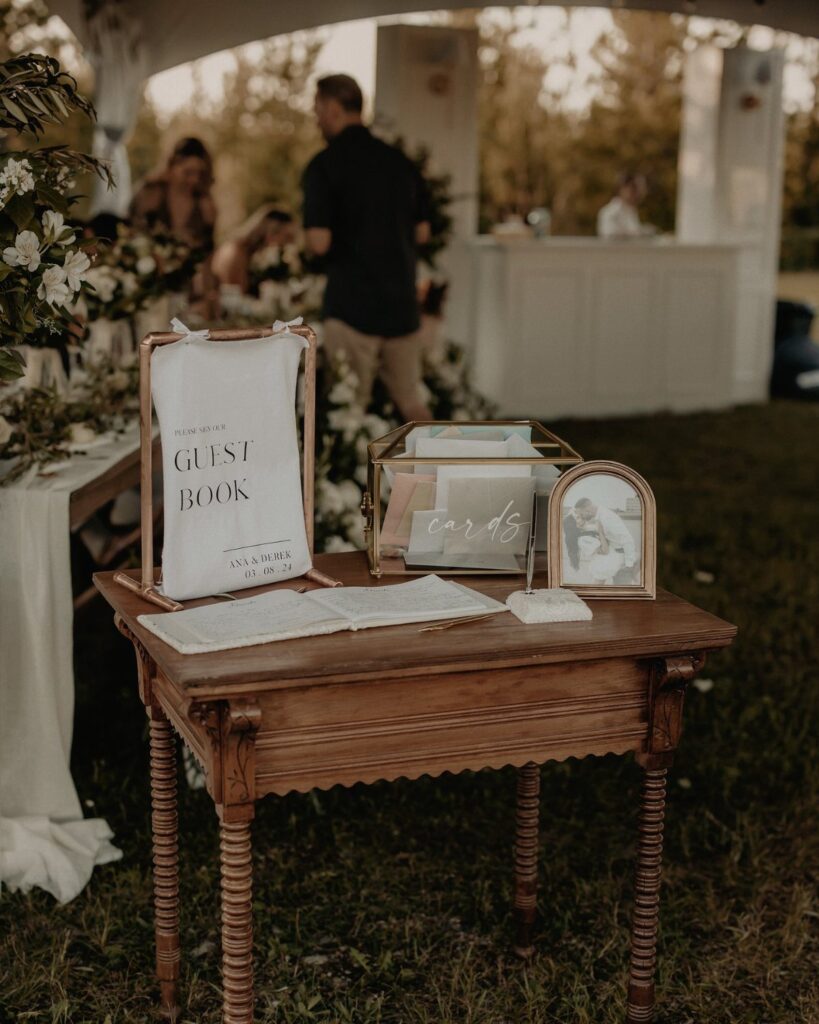 A wooden table set with a guest book, card box, and sign at an outdoor wedding.