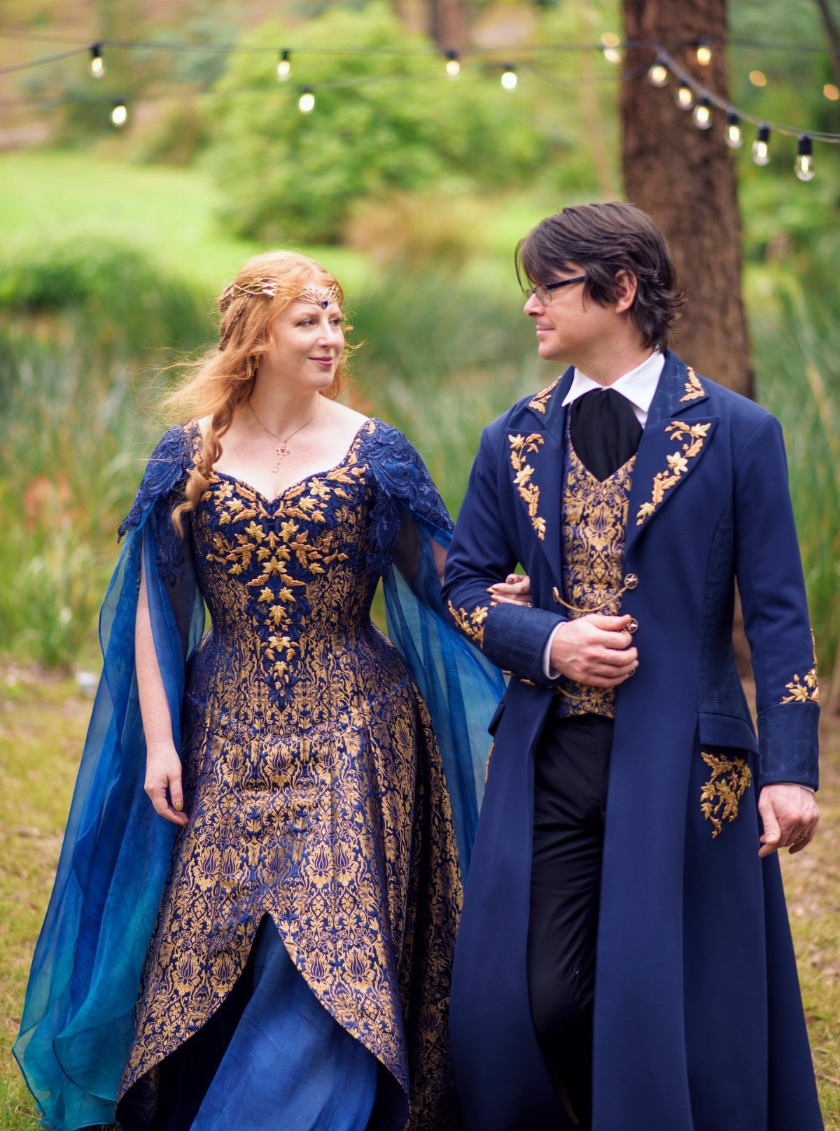 A couple in ornate blue and gold fantasy wedding attire walking through a green outdoor setting.