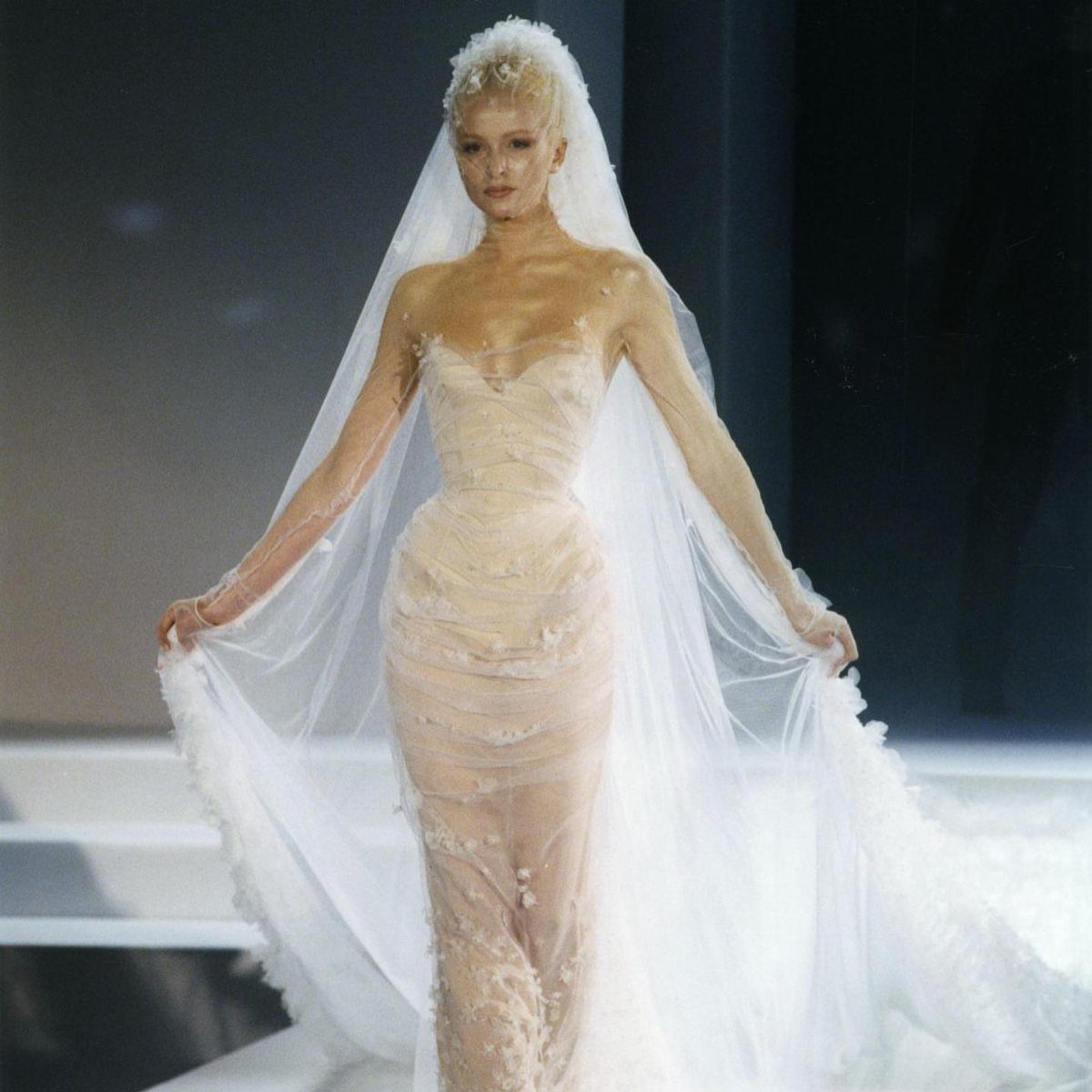 A model walking a runway in a sheer, textured white wedding dress and long veil.
