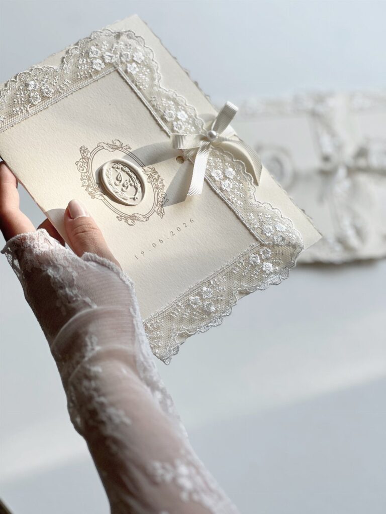 A hand in a lace sleeve holding a cream wedding invitation decorated with lace and a ribbon.